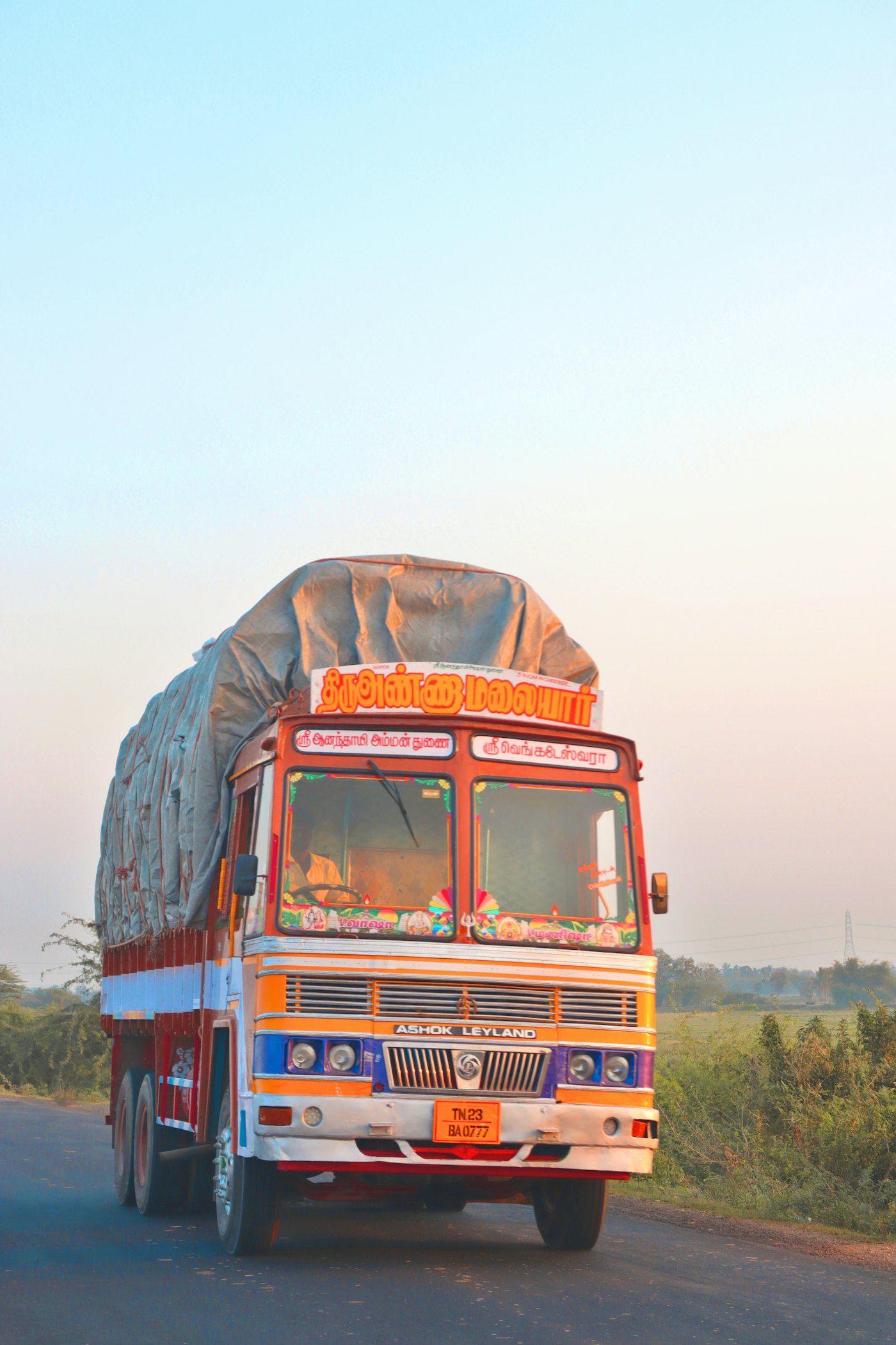 A loaded Indian goods truck, painted orange and blue, driving along a rural highway at sunrise