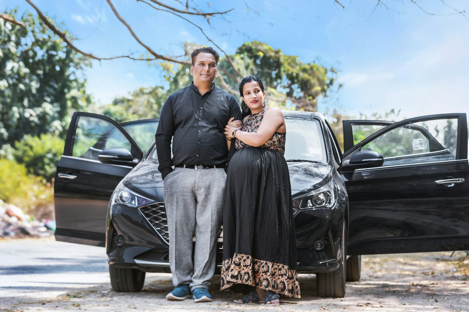A young couple, expecting a baby, standing beside their black Hyundai hatchback on a sunny day