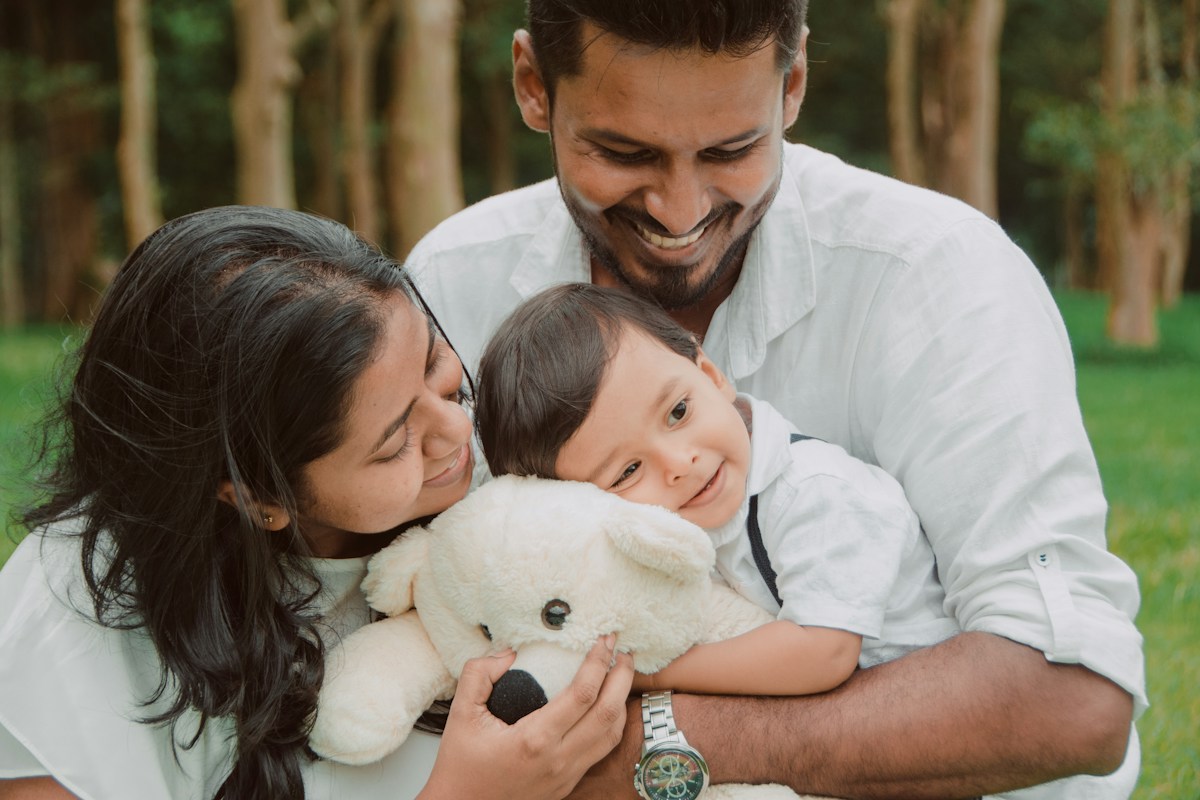 Smiling parents hugging their baby, who holds a teddy bear, in a sunny park