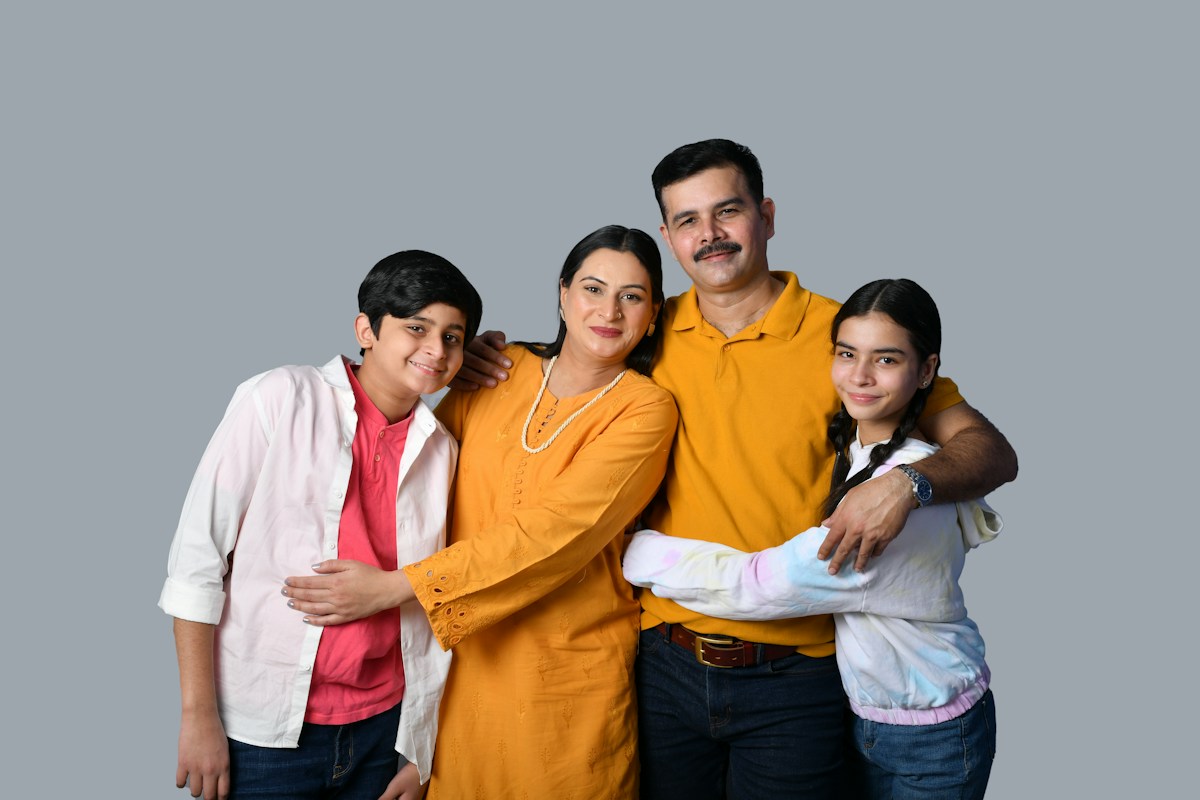 A smiling family of four, parents with their son and daughter, hugging against a grey background